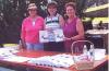 Three volunteers standing behind a table selling copies of the Our Lady of Good Council Parish History during the Skaro Pilgrimage